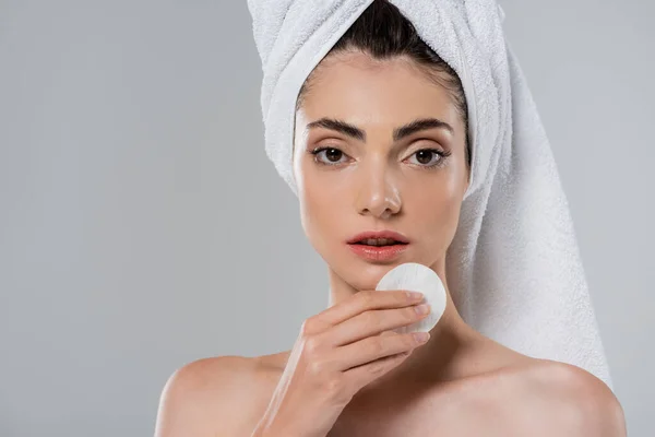 Jeune femme avec serviette sur la tête démaquillage avec tampon de coton isolé sur gris — Photo de stock