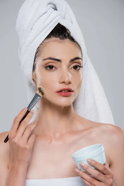 Mujer joven con toalla en la cabeza aplicando mascarilla con cepillo cosmético aislado en gris - foto de stock