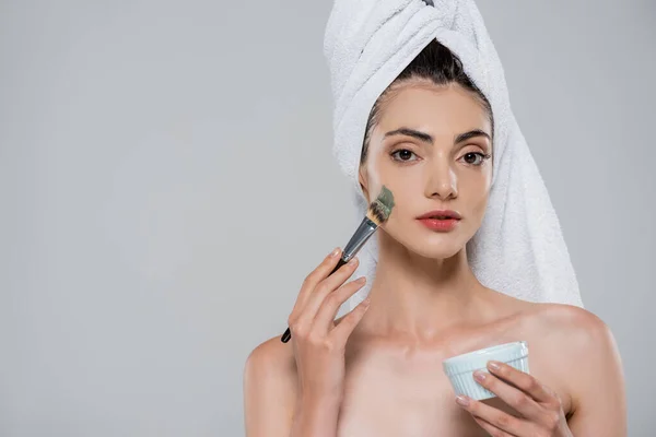 Young woman with towel on head applying green clay mask with cosmetic brush isolated on grey — Stock Photo