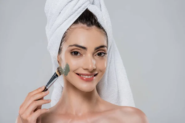 Cheerful young woman with towel on head applying green clay mask with cosmetic brush isolated on grey — Stock Photo