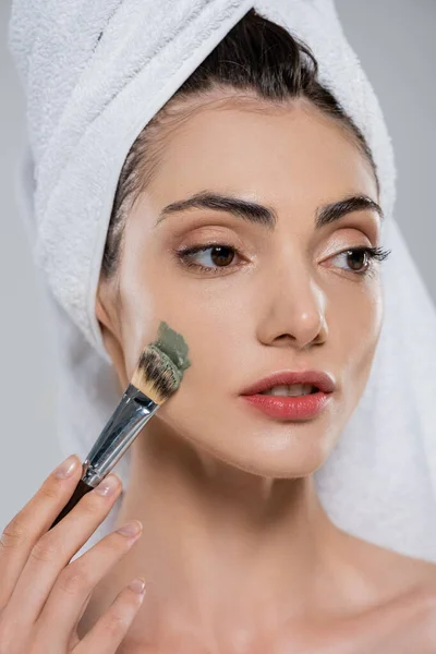 Junge Frau mit Handtuch auf dem Kopf Anwendung Tonmaske mit Kosmetikbürste isoliert auf grau — Stockfoto