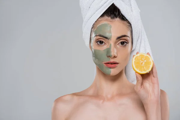 Young woman with towel on head and green clay mask on face holding half of orange isolated on grey — Stock Photo