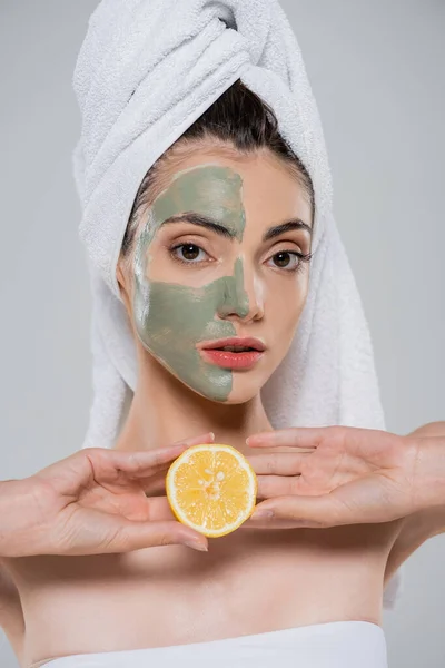 Mujer joven máscara de arcilla verde en la cara sosteniendo la mitad de naranja aislado en gris - foto de stock