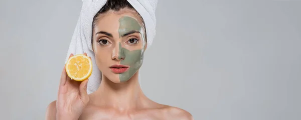 Mujer joven máscara de arcilla verde en la cara sosteniendo la mitad de naranja aislado en gris, bandera - foto de stock