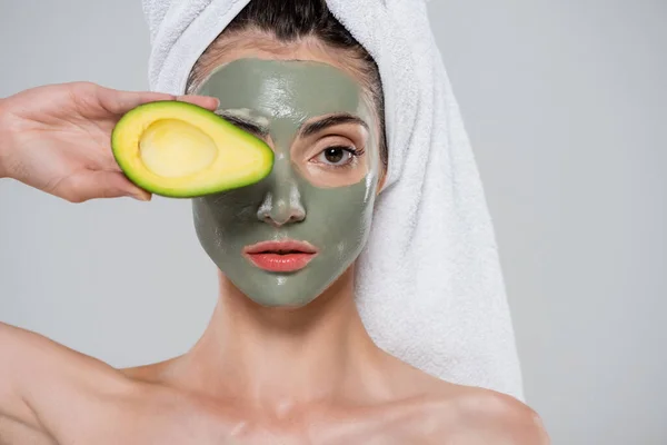 Junge Frau mit Handtuch auf dem Kopf und Tonmaske, die Auge mit Avocado auf grau isoliert — Stockfoto