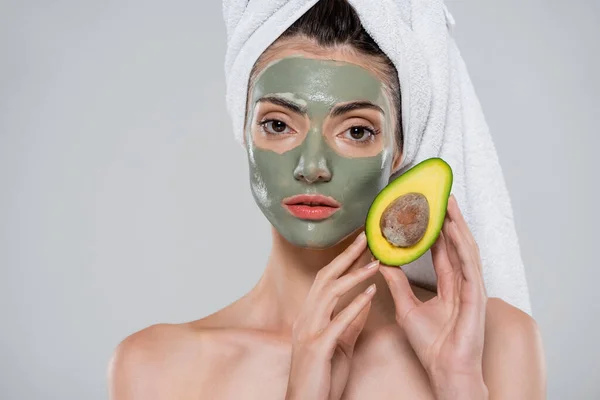 Jeune femme avec serviette sur la tête et masque d'argile verte tenant avocat mûr isolé sur gris — Photo de stock