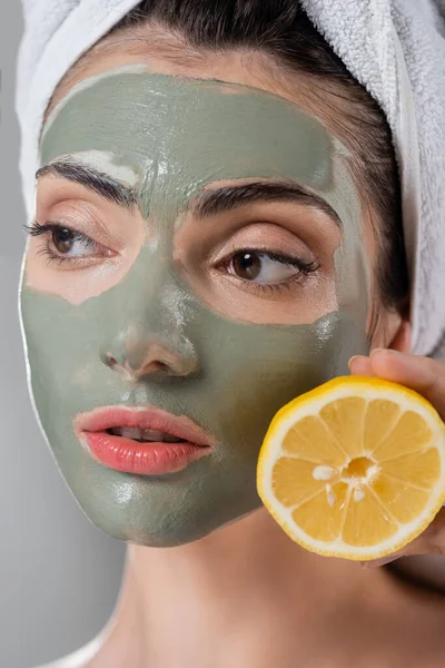 Primer plano de mujer joven con máscara de arcilla verde sosteniendo naranja madura medio aislado en gris - foto de stock
