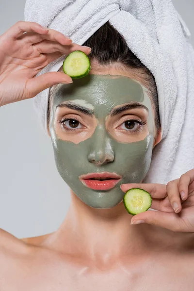 Jeune femme avec serviette sur la tête et masque d'argile verte sur le visage tenant concombre tranché isolé sur gris — Photo de stock