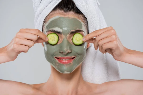 Felice giovane donna con maschera di argilla verde sul viso che copre gli occhi con cetriolo affettato isolato su grigio — Foto stock