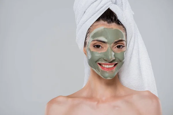 Радісна молода жінка з рушником на голові і зелену глиняну маску на обличчі ізольовано на сірому — стокове фото