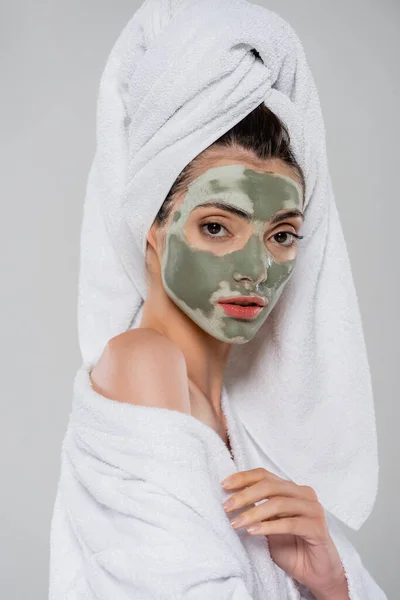 Young woman with towel on head and clay mask on face looking at camera isolated on grey — Stock Photo