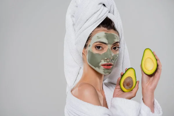 Молода жінка з рушником на голові і зеленою глиняною маскою, що тримає половинки авокадо ізольовані на сірому — стокове фото