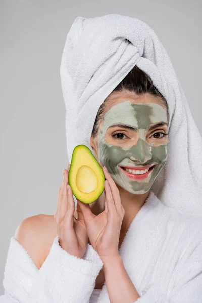 Allegra giovane donna con asciugamano sulla testa e maschera di argilla verde con avocado fresco isolato su grigio — Foto stock