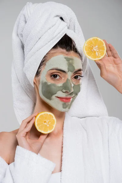 Lächelnde junge Frau mit grüner Lehmmaske, die reife orangefarbene Hälften isoliert auf grau hält — Stockfoto
