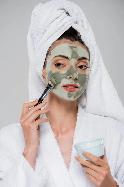 Young woman with towel on head applying clay mask with cosmetic brush while looking away isolated on grey — Stock Photo