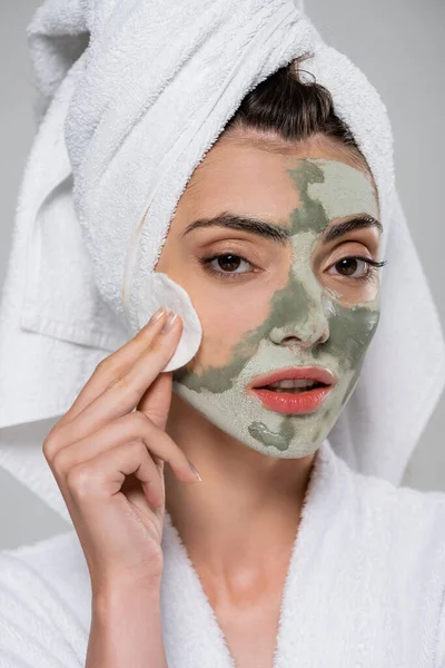 Young woman in bathrobe removing clay mask with cotton pad isolated on grey — Stock Photo