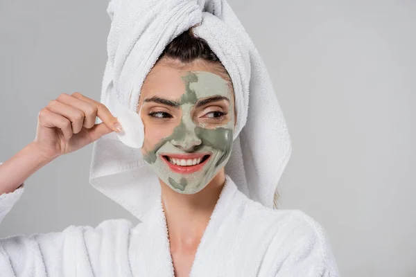 Happy young woman in bathrobe removing clay mask with cotton pad isolated on grey — Stock Photo