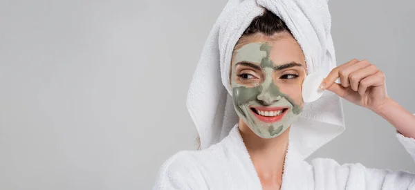 Happy young woman in bathrobe removing clay mask with cotton pad isolated on grey, banner — Stock Photo