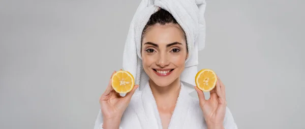 Cheerful young woman with towel on head holding halves of orange isolated on grey, banner — Stock Photo