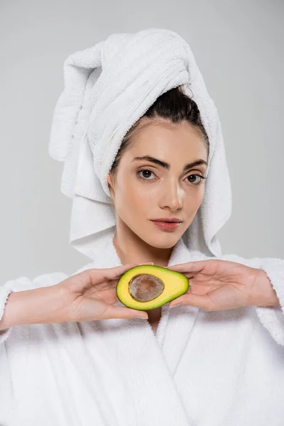 Mujer joven en albornoz sosteniendo la mitad del aguacate aislado en gris - foto de stock