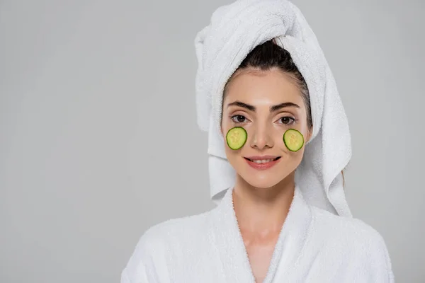 Giovane donna sorridente con asciugamano sulla testa e cetriolo affettato sul viso isolato sul grigio — Foto stock