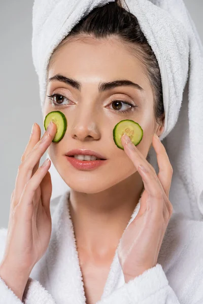 Mujer joven con toalla en la cabeza aplicando pepino en rodajas en la cara aislado en gris - foto de stock