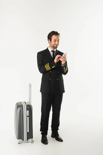 Giovane pilota utilizzando smartphone vicino alla valigia su sfondo bianco — Foto stock