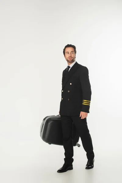 Pilot in uniform holding suitcase while walking on white background — Stock Photo
