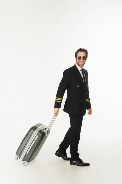Piloto sonriente en gafas de sol sosteniendo la maleta mientras camina sobre fondo blanco - foto de stock