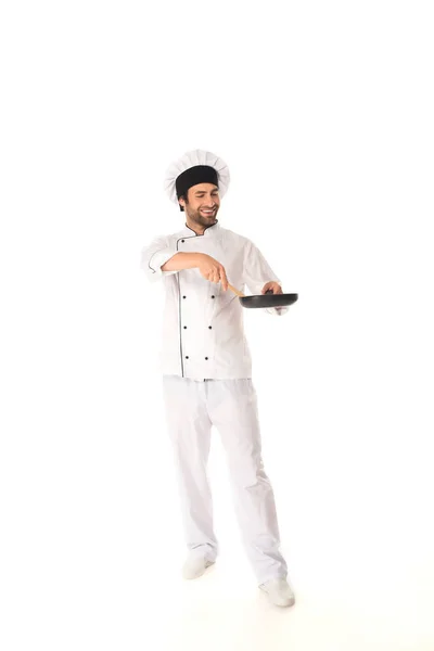 Longitud completa de chef sonriente sosteniendo sartén y espátula de madera sobre fondo blanco - foto de stock