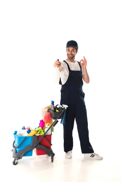 Sorrindo limpador segurando detergente e mostrando ok perto carrinho no fundo branco — Fotografia de Stock