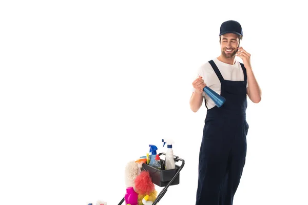 Smiling cleaner talking on smartphone and holding detergent near cart isolated on white — Stock Photo