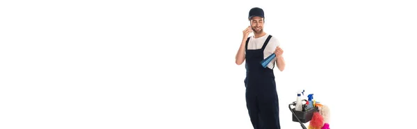 Cleaner in uniform talking on smartphone and holding detergent near cart isolated on white, banner — Stock Photo