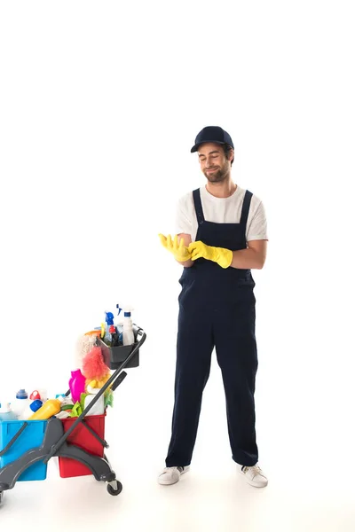 Nettoyant souriant portant des gants en caoutchouc près du chariot sur fond blanc — Photo de stock