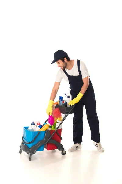 Side view of smiling cleaner standing near cart with detergents on white background — Stock Photo