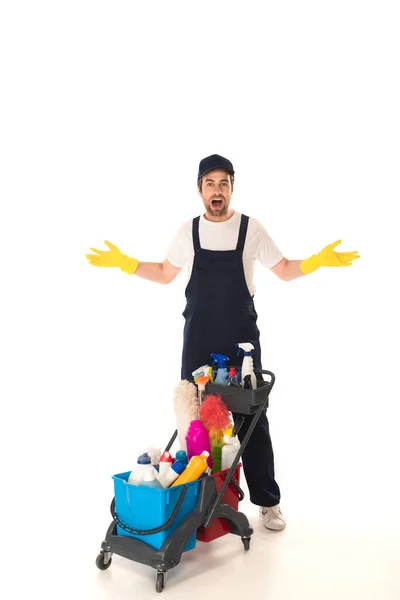 Amazed cleaner in rubber gloves looking at camera near cart on white background — Stock Photo