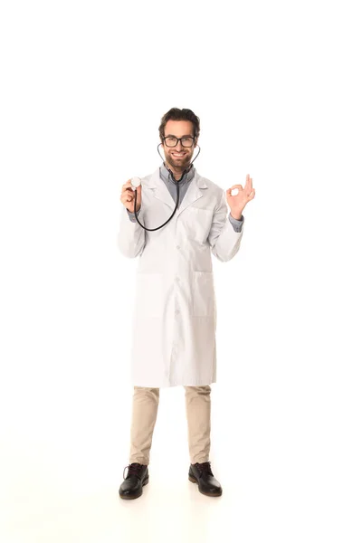 Médico sonriente sosteniendo estetoscopio y mostrando un gesto aceptable sobre fondo blanco - foto de stock