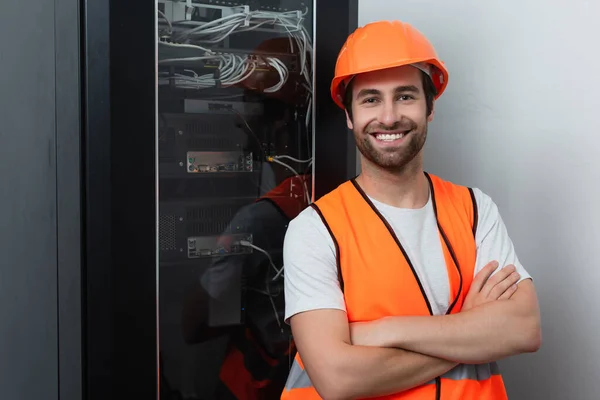 Positive workman in safety vest standing with crossed arms near switchboard — Stock Photo