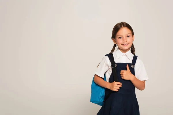 Happy schoolgirl with blue backpack showing thumb up isolated on grey — Stock Photo