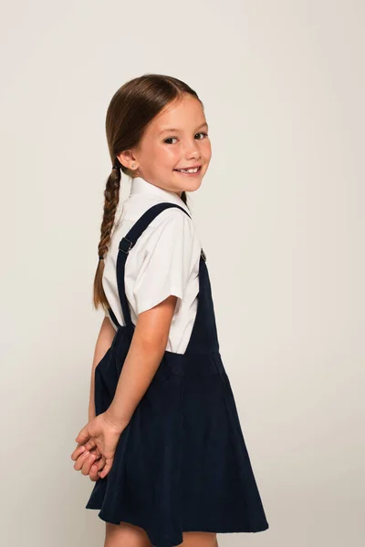 Positive schoolchild standing with hands behind back and looking at camera isolated on grey — Stock Photo