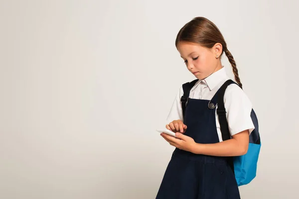 Menina em uniforme escolar mensagens no telefone celular isolado no cinza — Fotografia de Stock