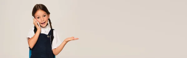 Colegial positivo apuntando con la mano mientras habla en el teléfono inteligente aislado en gris, bandera - foto de stock