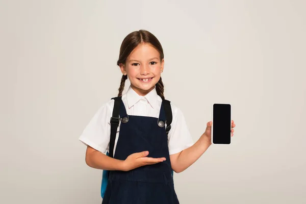 Criança da escola sorrindo apontando para o smartphone com tela em branco isolada em cinza — Fotografia de Stock