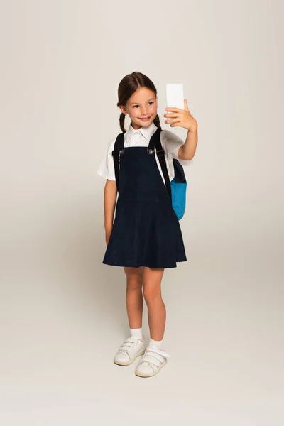 Ganzkörperansicht des Mädchens in Schuluniform, das ein Selfie mit dem Handy auf grau macht — Stockfoto