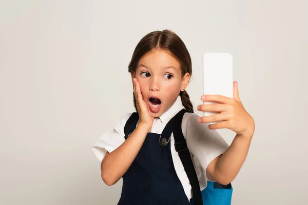 Asombrado escolar tocando la cara mientras toma selfie en el teléfono celular aislado en gris - foto de stock