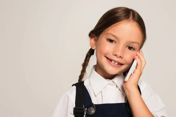 Glückliches Schulmädchen, das in die Kamera schaut, während es auf dem Smartphone spricht, isoliert auf grau — Stockfoto