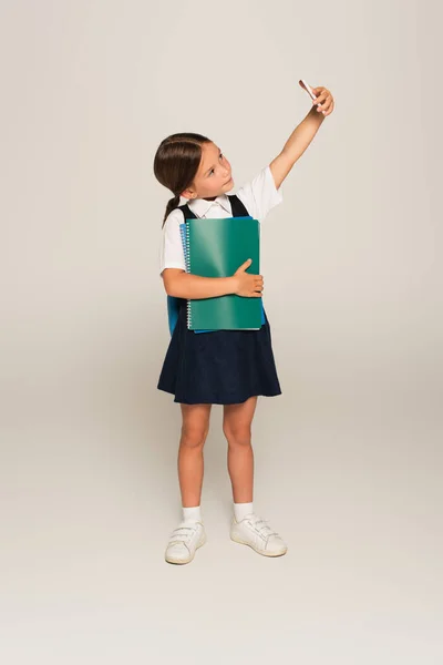 Full length view of schoolgirl with copy books taking selfie on cellphone on grey — Stock Photo