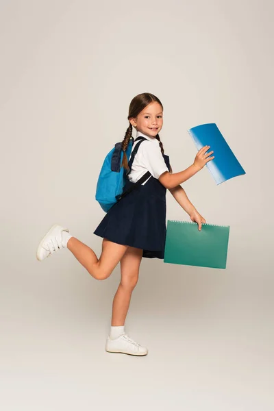 Ganzkörperansicht des fröhlichen Schulkindes, das mit Notizbüchern auf grau läuft — Stockfoto