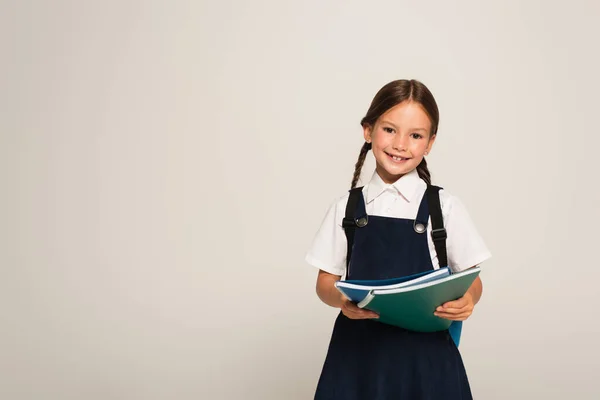 Positives Schulkind lächelt in die Kamera und hält Hefte isoliert auf grau — Stockfoto