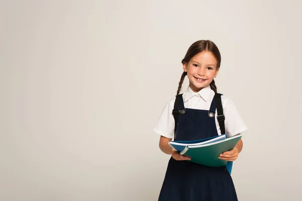 Colegiala positiva sonriendo a la cámara mientras sostiene cuadernos aislados en gris - foto de stock